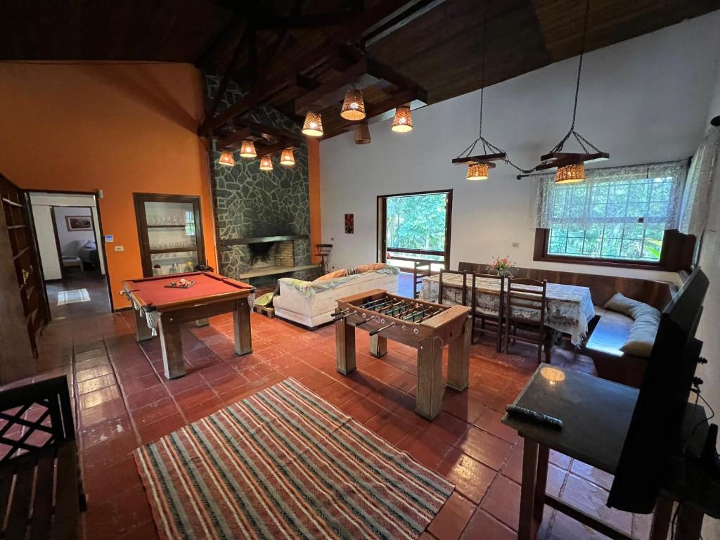 Imagem de um espaço interno aconchegante da Casa Penedo, apresentando uma sala de lazer com mesa de sinuca, totó, uma lareira revestida em pedra e um ambiente iluminado com luminárias rústicas pendentes. O espaço possui piso de cerâmica avermelhada, janelas amplas que oferecem vista para a vegetação externa e áreas de descanso com sofás e mesa de jantar.