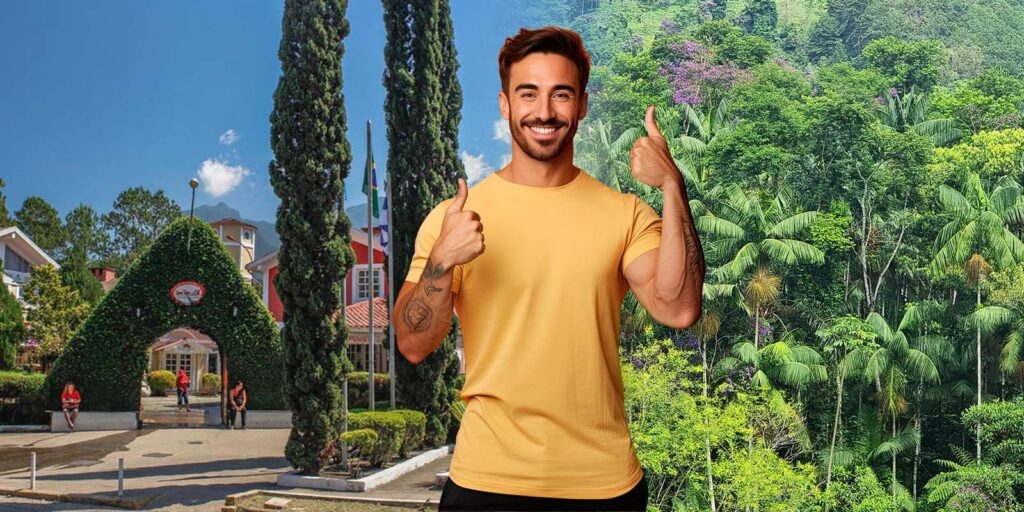 Imagem de um homem sorridente vestindo uma camiseta amarela, com os polegares levantados, em frente à entrada arborizada da Pequena Finlândia em Penedo, com a natureza exuberante da Serra da Mantiqueira ao fundo.