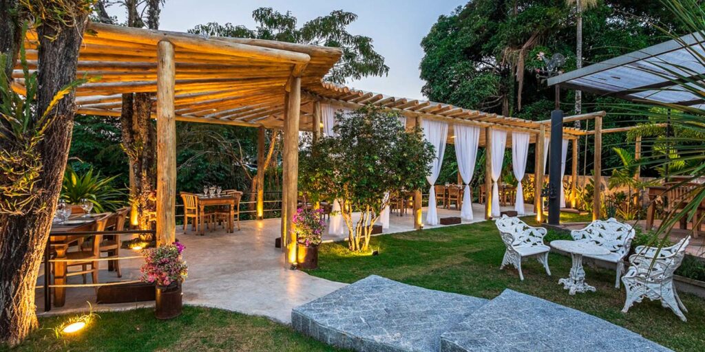 Área externa do Restaurante Botegare Penedo ao entardecer, com pergolado de madeira, cortinas brancas, mesas elegantemente postas e iluminação aconchegante em meio à natureza.