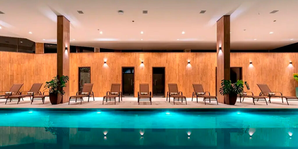 Piscina aquecida coberta do Vert Hotel Spa Penedo, cercada por espreguiçadeiras e uma elegante parede de madeira com iluminação aconchegante.